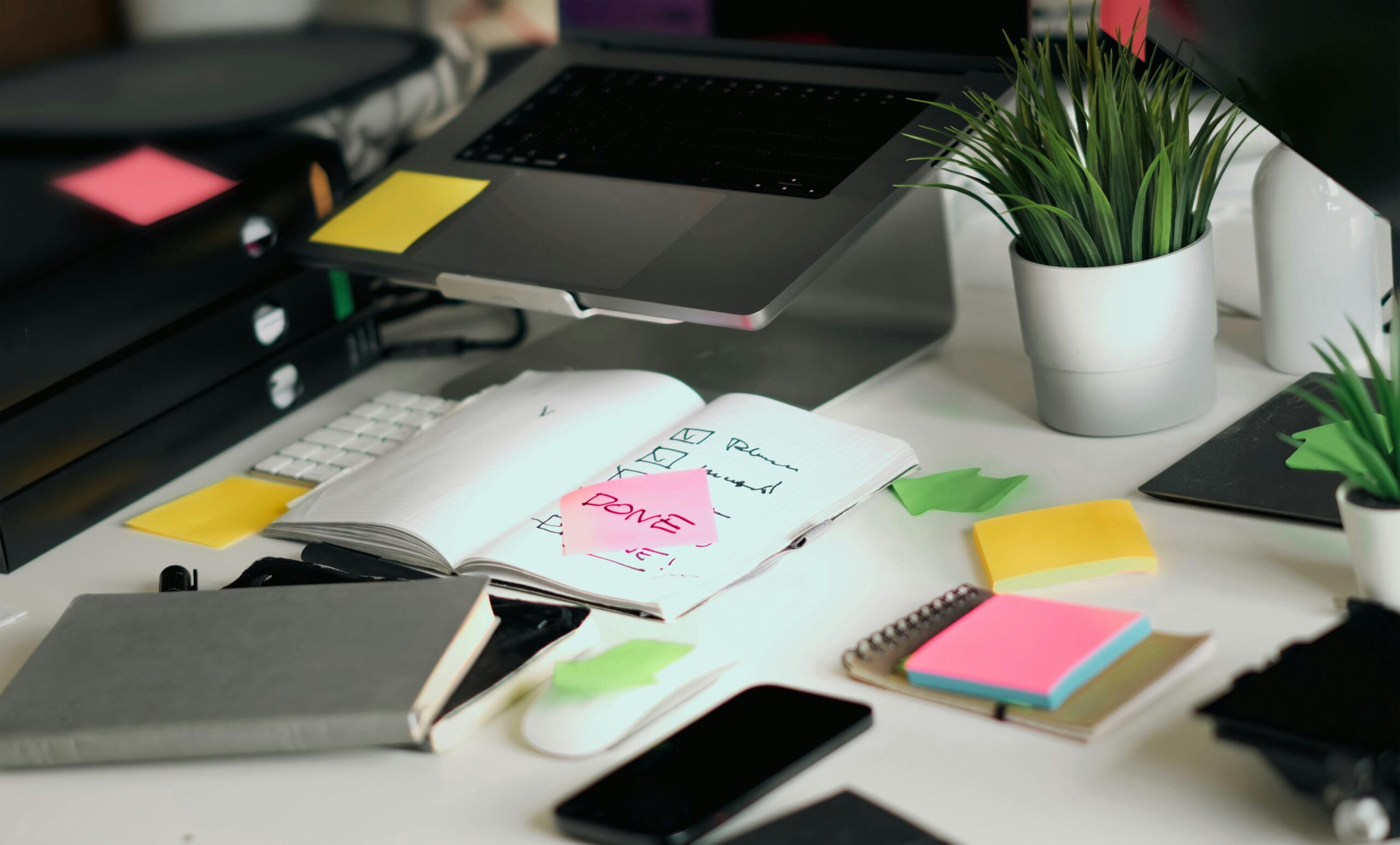 A cluttered office desk featuring a laptop, notebooks, and checklists amid colorful sticky notes.